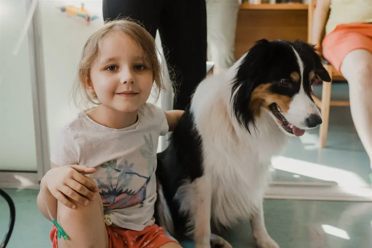 Uśmiechnięta dziewczynka z wenflonem siedzi obok psa terapeutycznego. Psy do dogoterapii przynoszą radość i wsparcie.